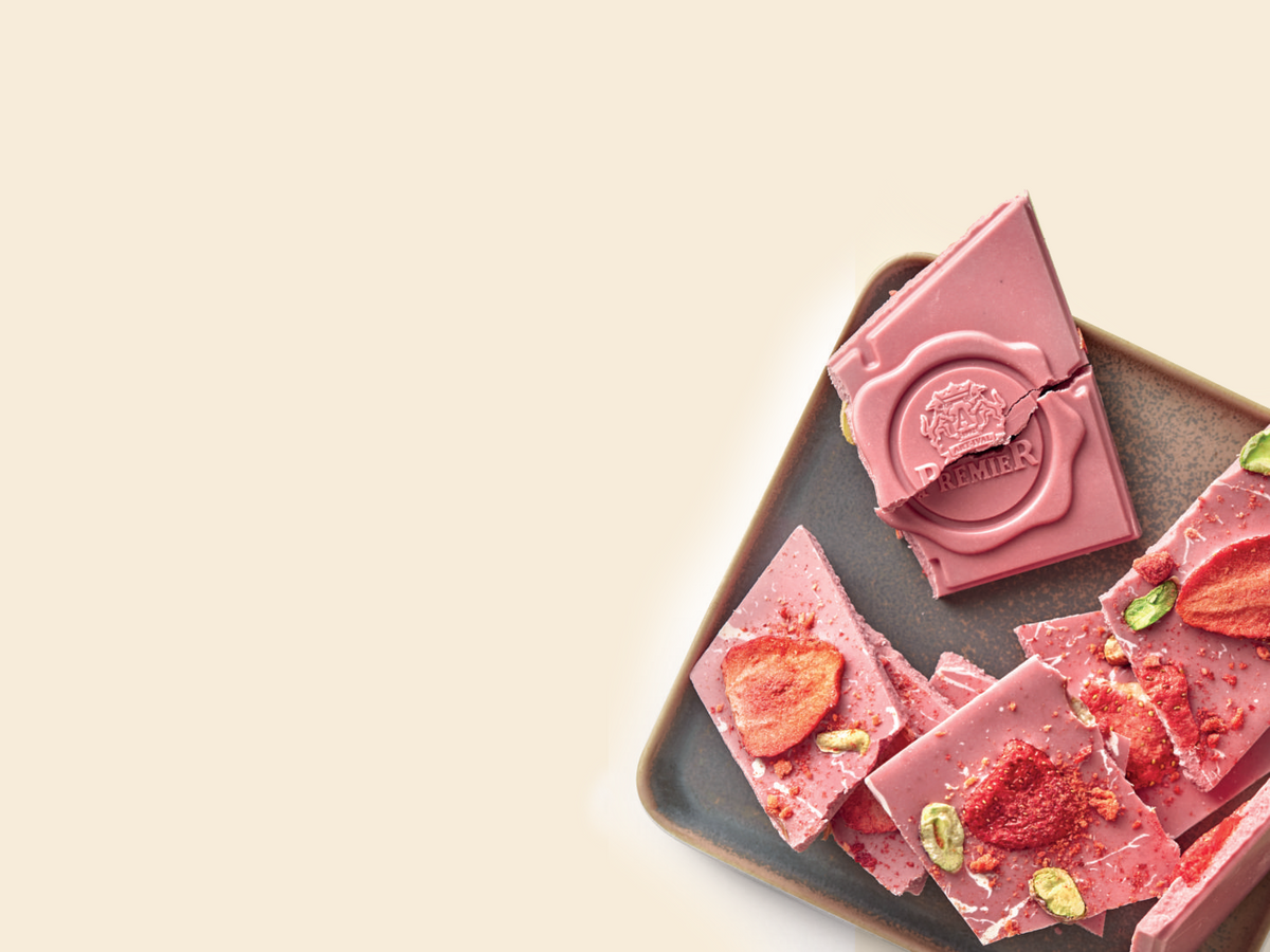 Ruby chocolate bark pieces on a dark square tray, including a broken section showing the embossed 'Chocolat des Jardins' logo, dried strawberry slices, and green pistachio nuts, set against a warm cream background.